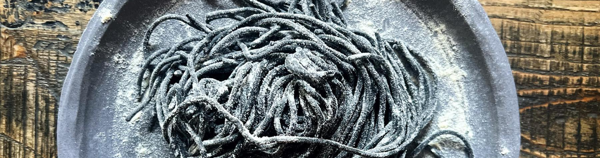 Na obrazie widzimy talerz z czarnym makaronem, który posypany jest mąką, co nadaje mu delikatnie pudrowanego wyglądu. Całość jest prezentowana na drewnianej powierzchni.