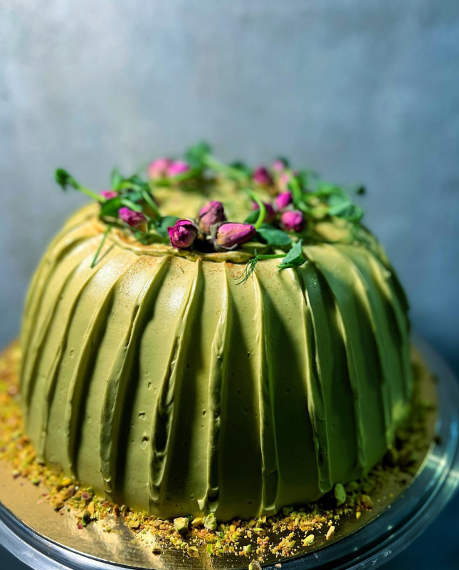 Na obrazie widzimy pięknie udekorowany tort o gładkim, zielonym lukrze, z delikatnymi pionowymi żłobieniami na bokach. Na wierzchu tortu znajdują się różowe pąki róż oraz zielone listki, starannie rozmieszczone w formie dekoracyjnej kompozycji. U podstawy ciasta, na okrągłym talerzu, rozsypane są pokruszone orzechy lub okruszki, które dodają całości tekstury.