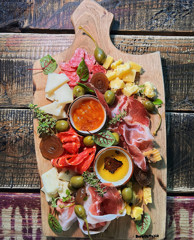 Na obrazie widzimy pięknie zaaranżowaną deskę przekąsek, idealną na przyjęcie lub spotkanie towarzyskie. Na drewnianej desce znajdują się różne rodzaje serów, pokrojone w kostki i kliny, a także cienko krojone wędliny. Widzimy również oliwki oraz dwie małe miseczki z sosami. Wszystko ozdobione jest świeżymi ziołami i liśćmi, co nadaje całości świeżego i eleganckiego wyglądu.