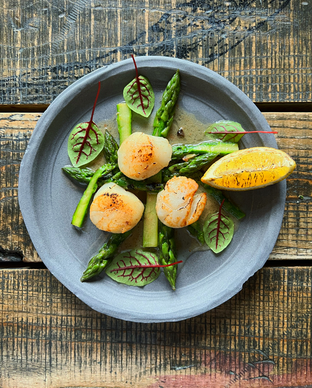 Na zdjęciu widzimy danie o eleganckiej prezentacji, które spoczywa na drewnianym stole, tworząc rustykalne i stylowe tło. Na talerzu znajdują się podsmażone przegrzebki, zielone szparagi oraz listki z czerwonymi żyłkami. Na talerzu znajduje się również kawałek grillowanej cytryny, co sugeruje, że danie może być doprawione jej kwaskowym smakiem.