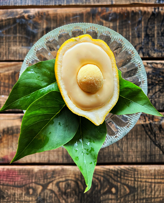 Na obrazie widzimy połówkę cytryny, która została wydrążona i wypełniona kremową substancją. Na wierzchu znajduje się mały okrągły element, prawdopodobnie owoc lub cukierek, który stanowi dekorację. Cytryna umieszczona jest na szklanym talerzyku, a wokół niej rozłożone są zielone liście.