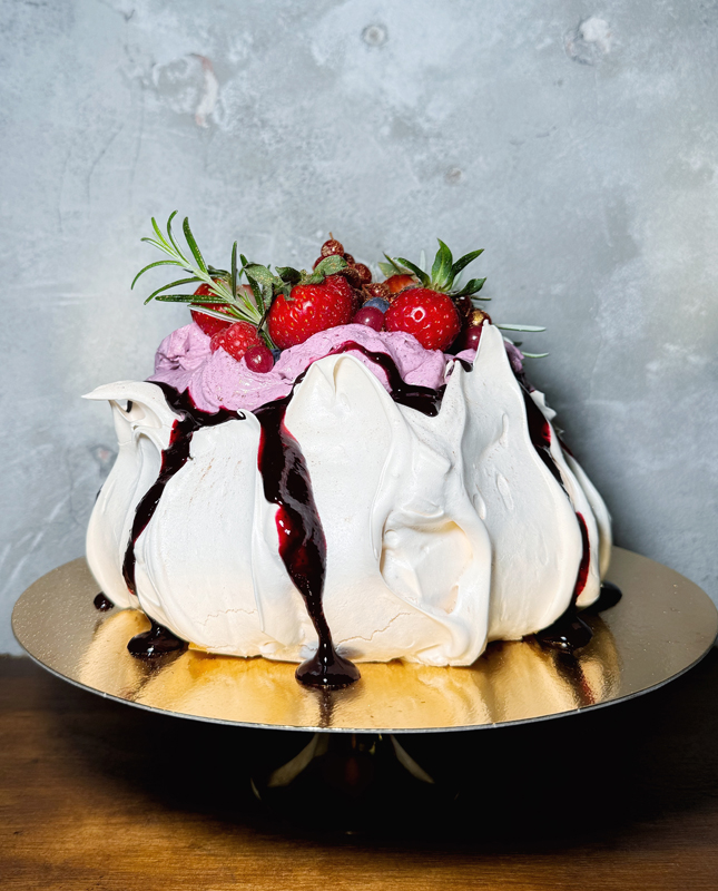 Na obrazie widzimy przepysznie udekorowany deser, najprawdopodobniej pavlovą lub tort bezowy. Ma nieregularną, puszystą strukturę, charakterystyczną dla bezy, a wierzch pokryty jest obfitą warstwą fioletowego kremu lub musu. Na szczycie znajdują się świeże truskawki, które dodają intensywnego koloru, a także gałązki rozmarynu.