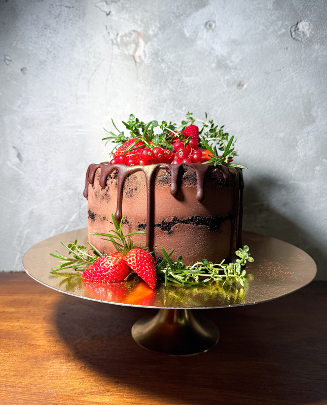 Na obrazie widzimy pięknie udekorowany tort czekoladowy, ustawiony na złotej paterze. Tort pokryty jest bogatą polewą czekoladową, która spływa po bokach, dodając mu wyrafinowanego charakteru. Na wierzchu tortu umieszczone są świeże truskawki, czerwone porzeczki i dekoracyjne gałązki zieleni.