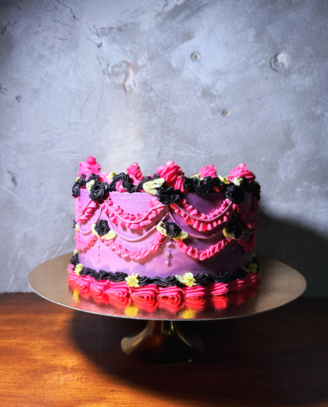 Na obrazie widzimy imponujący tort na metalowej paterze. Tort ma fioletową bazę z kunsztownymi dekoracjami w kolorach różowym, czarnym i żółtym. Dekoracje obejmują wiry, rozety oraz drapowane wzory, które nadają całości świąteczny i bogaty charakter. Tło stanowi teksturowana, szara ściana, a tort umieszczono na drewnianej powierzchni.