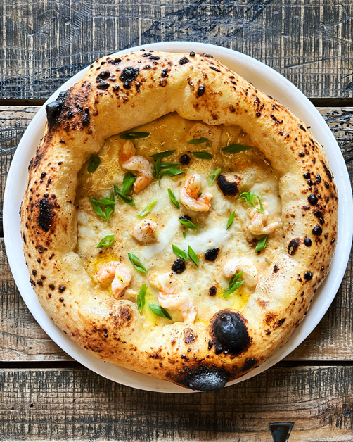 Na obrazie widzimy pizzę na białym talerzu, która wyróżnia się grubym, przypieczonym na czarno brzegiem. Pizza ma biały sos jako bazę, a na wierzchu znajdują się krewetki i posypka z zielonej cebulki, które dodają koloru i świeżości.