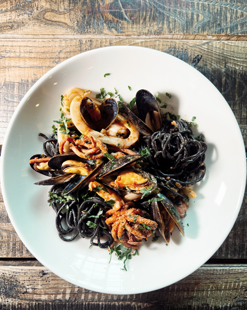 Na obrazie widzimy apetyczne danie - talerz makaronu z owocami morza. Makaron ma czarny kolor. Wśród dodatków znajdują się mule i kawałki kalmara. Danie udekorowane jest drobno posiekanymi ziołami, które dodają świeżości i koloru. Wszystko podane jest na białym talerzu, który kontrastuje z rustykalnym, drewnianym stołem w tle.