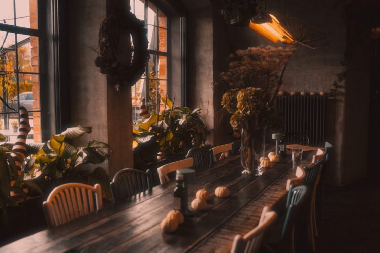 Na obrazie widzimy przytulną restaurację, w której centralnym punktem jest długi drewniany stół otoczony krzesłami. Stół jest udekorowany małymi dyniami oraz wazonem z suszonymi kwiatami, co podkreśla jesienny klimat wnętrza. Po lewej stronie znajdują się duże okna, które wpuszczają naturalne światło i zapewniają widok na zewnątrz. Okna ozdobione są wieńcami, a wokół nich rozstawione są różne rośliny, dodając pomieszczeniu ciepła i naturalnego uroku. W centralnej części pomieszczenia wisi lampa z ciepłym światłem, które tworzy przytulną atmosferę.