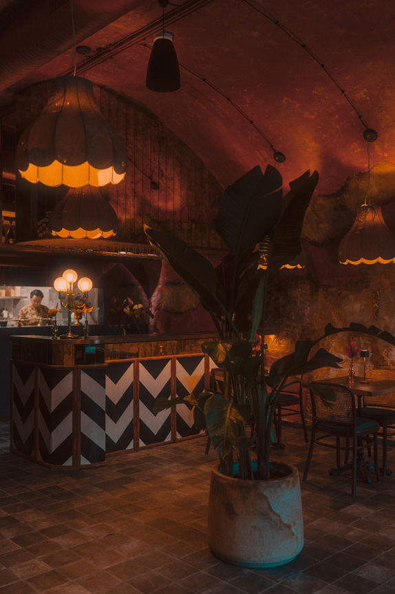 Na obrazie widzimy stylowe wnętrze restauracji o ciepłym, nastrojowym oświetleniu. Wystrój wnętrza jest eklektyczny i przytulny – sufit ma zakrzywioną, przypominającą jaskinię strukturę, z której zwisają designerskie lampy o ozdobnych, falistych krawędziach, emitujące delikatne, żółte światło. W centralnej części znajduje się bar o wykończeniu w modny wzór w jodełkę, co przyciąga wzrok. Na pierwszym planie widzimy duży, zielony doniczkowy kwiat.