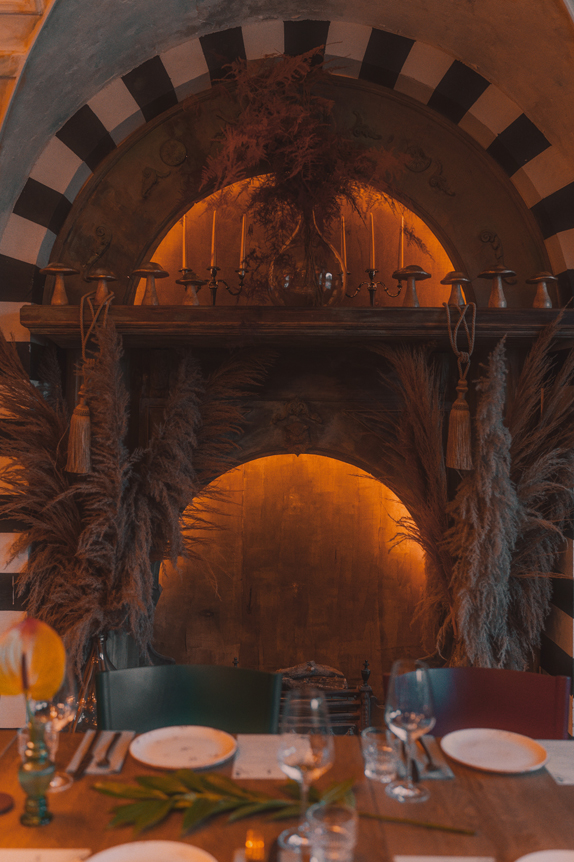 Na obrazie widzimy elegancko urządzone miejsce jadalne z rustykalnym akcentem, koncentrujące się na wnęce w ścianie. Wnęka ma półokrągły łuk ozdobiony naprzemiennymi czarnymi i białymi paskami, co przyciąga uwagę i dodaje artystycznego charakteru. Na półce umieszczone są różne dekoracyjne przedmioty, takie jak świeczniki oraz wazon z suszonymi kwiatami, podkreślające przytulną atmosferę. Poniżej półki, po obu stronach, znajdują się duże suszone rośliny, które wzmacniają naturalny i ciepły klimat wnętrza. W tle widać stół jadalniany nakryty talerzami, szklankami i sztućcami, co wskazuje na przygotowanie przestrzeni do posiłku.