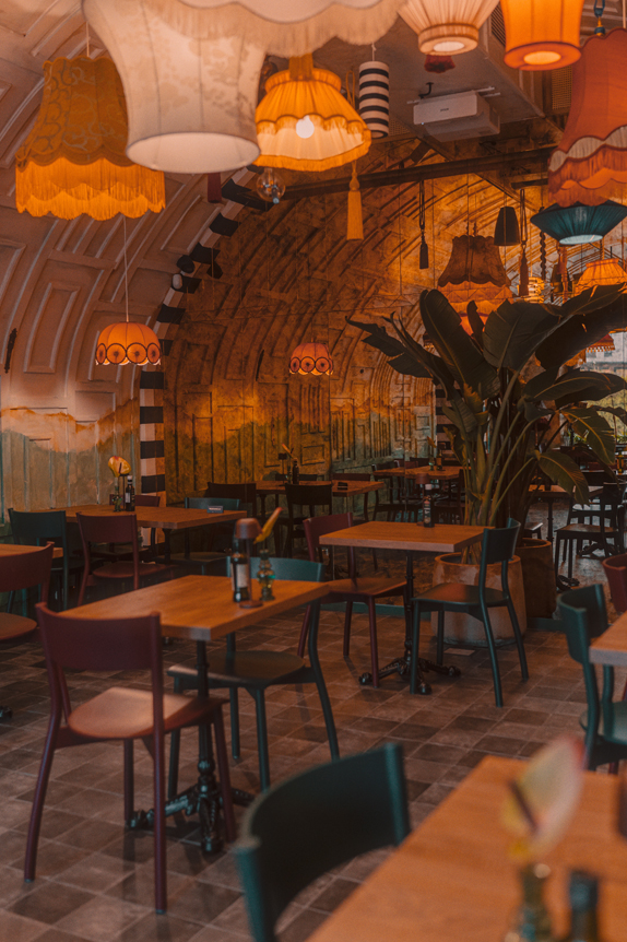 Na obrazie widzimy wnętrze przytulnej i eklektycznej restauracji. Wiszące lampy w stylu vintage są różnokolorowe, co nadaje przestrzeni żywego charakteru i ciepłej, zachęcającej atmosfery. Stoły i krzesła są różnorodne i niedopasowane, co podkreśla artystyczny i swobodny styl wnętrza. W centrum pomieszczenia znajduje się duża roślina doniczkowa.