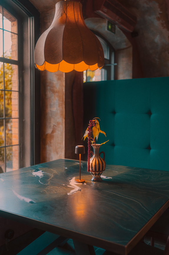 Na obrazie widzimy przytulne wnętrze z centralnym punktem w postaci stołu. Na stole znajduje się dekoracyjny wazon z rośliną, a obok stoi mała, nowoczesna lampka, która dodaje przestrzeni uroku. Nad stołem wisi lampa o falistym, ozdobnym kloszu, emitująca ciepłe, żółte światło, tworząc nastrojową atmosferę. W tle widoczna jest turkusowa, pikowana ławka oraz teksturowana ściana.