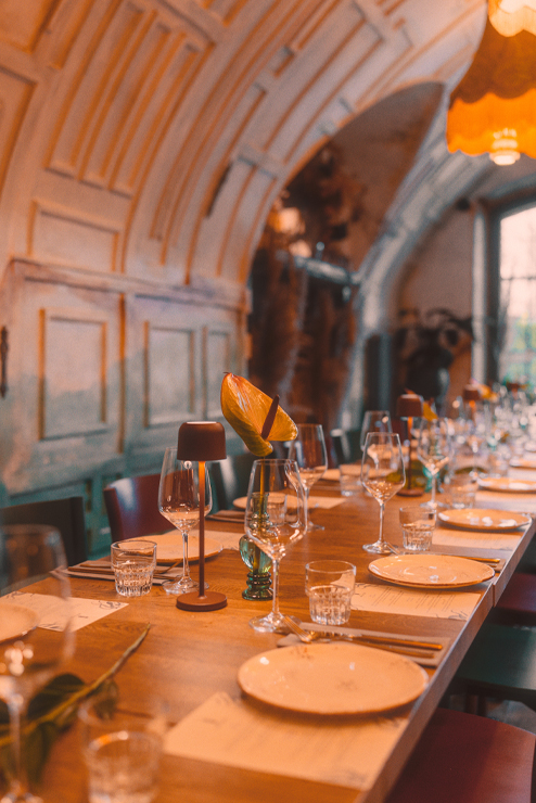 Na obrazie widzimy długi stół jadalniany, przygotowany na posiłek w eleganckiej restauracji. Stół jest starannie nakryty – znajdują się na nim talerze, sztućce, kieliszki do wina, szklanki oraz serwetki, wszystko estetycznie rozmieszczone. Dekorację stanowi mały wazon z kwiatami oraz świecznik. Oświetlenie jest ciepłe, co tworzy zachęcającą atmosferę.