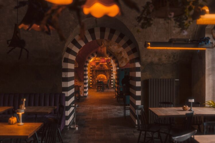 Obraz przedstawia wnętrze restauracji o przytulnej atmosferze. Wyjątkowym elementem są łukowe przejścia ozdobione czarno-białymi pasami, które tworzą efekt tunelu, przyciągając wzrok w głąb pomieszczenia. Oświetlenie jest ciepłe, o pomarańczowej barwie. W przestrzeni rozmieszczone są stoły i krzesła w uporządkowany sposób, a na stołach zauważalne są małe dekoracje, takie jak dynie, co sugeruje sezonowy lub świąteczny motyw.