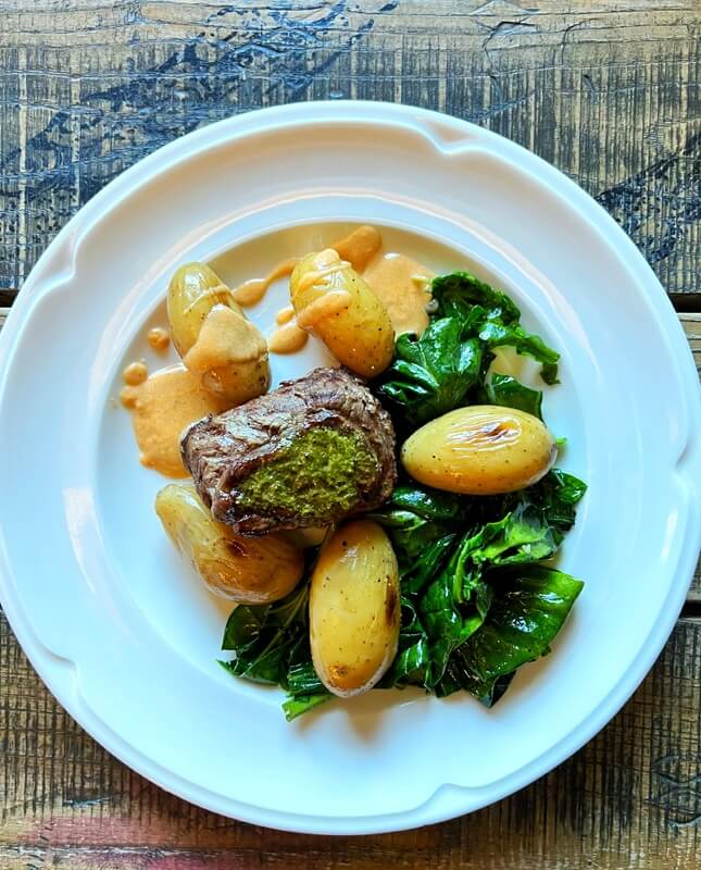 Na zdjęciu znajduje się podane danie - głównym elementem jest kawałek mięsa pokryty zielonym ziołowym sosem. Otaczają go małe pieczone ziemniaczki, aksamitny kremowy sos oraz porcja duszonych zielonych warzyw, prawdopodobnie szpinaku lub jarmużu. Całość zaserwowana jest na eleganckim białym talerzu, który stoi na drewnianym, rustykalnym stole.