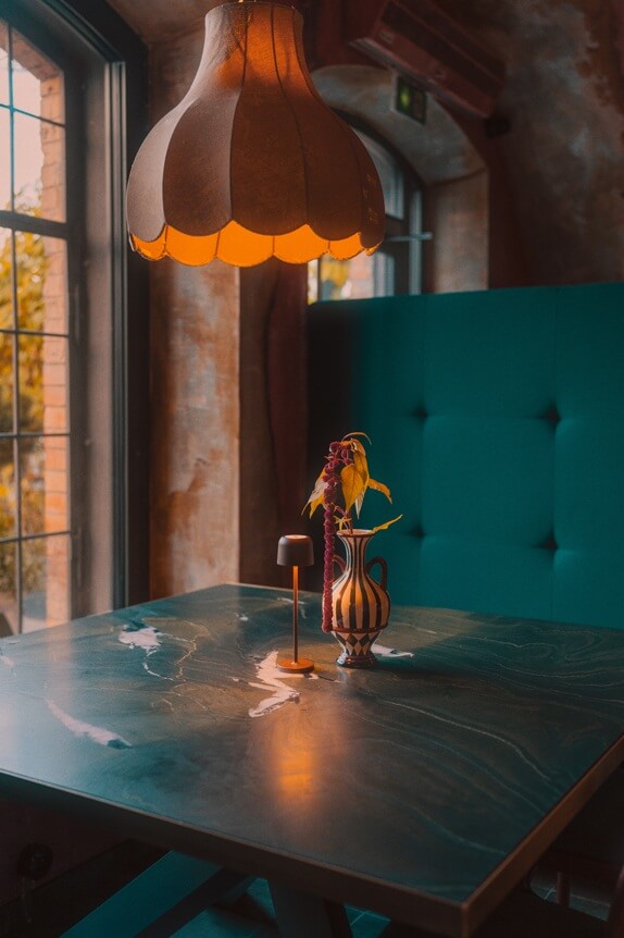 Na zdjęciu widzimy przytulny, starannie zaaranżowany kącik w restauracji. Centralnie znajduje się drewniany stół z małą lampką i wazonem z zieloną rośliną. Nad stołem wisi duża, ozdobna lampa, emitująca ciepłe, żółte światło. Z lewej strony zdjęcia znajduje się okno, przez które wpada naturalne światło, a w tle widać turkusową, tapicerowaną ławkę.
