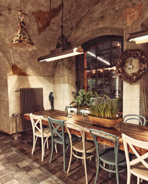 Na zdjęciu widzimy klimatyczne wnętrze restauracji, w której centralnym punktem jest długi, drewniany stół otoczony krzesłami w różnych kolorach i stylach. Ściany z surowego betonu, duże łukowe okno i wiszące lampy tworzą industrialno-rustykalny klimat, wzbogacony o detale vintage. Na stole znajduje się donica z zieloną rośliną, a w tle widoczna jest rzeźba przy grzejniku.